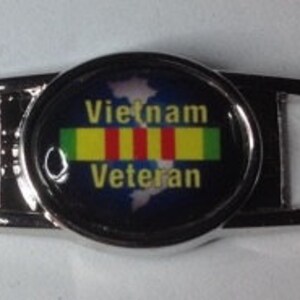 May include: Silver shoe charm with a blue background and a white outline of a map of Vietnam. The charm has the text "Vietnam Veteran" and a yellow, green, and red ribbon.