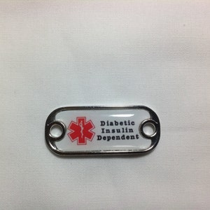 Puede incluir: Una etiqueta de metal plateado con fondo blanco y un símbolo médico rojo. La etiqueta dice "Diabetic Insulin Dependent".