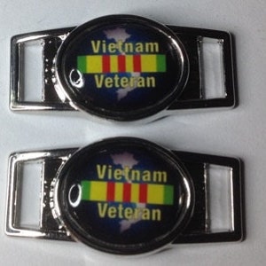 Puede incluir: Dos abalorios de zapatos plateados con fondo negro y un mapa del mundo. Los abalorios presentan una cinta verde, amarilla y roja con el texto "Vietnam Veteran".