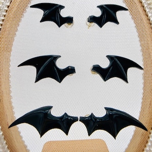 May include: A set of four black bat wing earrings. The earrings are displayed on a white background with a brown and white decorative frame.