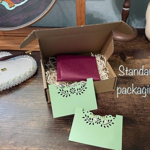 May include: A brown cardboard box with a burgundy tissue paper envelope inside. The box is filled with shredded paper and contains two green cards with a cut-out design. The text "Standard packaging" is visible on the right side of the image.