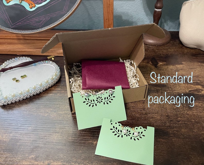 May include: A brown cardboard box with a burgundy tissue paper envelope inside. The box is filled with shredded paper and contains two green cards with a cut-out design. The text "Standard packaging" is visible on the right side of the image.