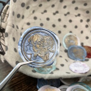 May include: A blue wax seal with a gold design of a woman's face and a floral pattern. The seal is being held by tweezers and is surrounded by other wax seals.