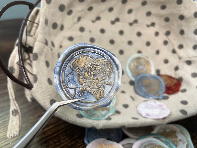 May include: A blue wax seal with a gold design of a woman's face and a floral pattern. The seal is being held by tweezers and is surrounded by other wax seals.