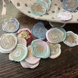 May include: A collection of round, decorative wax seals with various designs and colors, including gold, blue, pink, and white. The seals are arranged on a dark wooden surface.