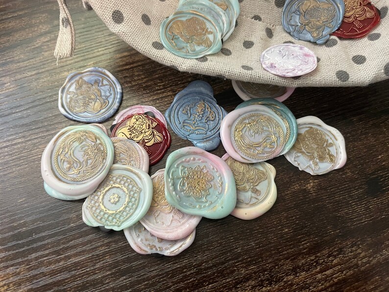 May include: A collection of round, decorative wax seals with various designs and colors, including gold, blue, pink, and white. The seals are arranged on a dark wooden surface.