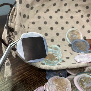 May include: A close-up of a silver-colored tweezers holding a round, blue and silver wax seal. The seal is blank and has a square shape in the center. The seal is surrounded by a variety of colorful wax seals in a basket.