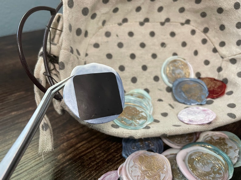 May include: A close-up of a silver-colored tweezers holding a round, blue and silver wax seal. The seal is blank and has a square shape in the center. The seal is surrounded by a variety of colorful wax seals in a basket.