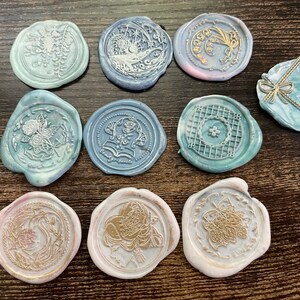 May include: Ten round wax seals with various designs in gold and white on a blue and white background. The designs include flowers, a moon, a star, a geometric pattern, and a butterfly.