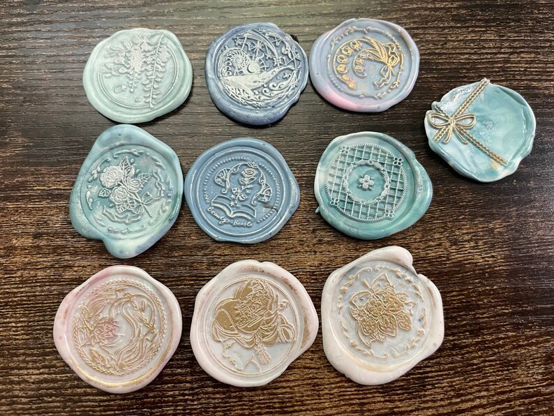 May include: Ten round wax seals with various designs in gold and white on a blue and white background. The designs include flowers, a moon, a star, a geometric pattern, and a butterfly.