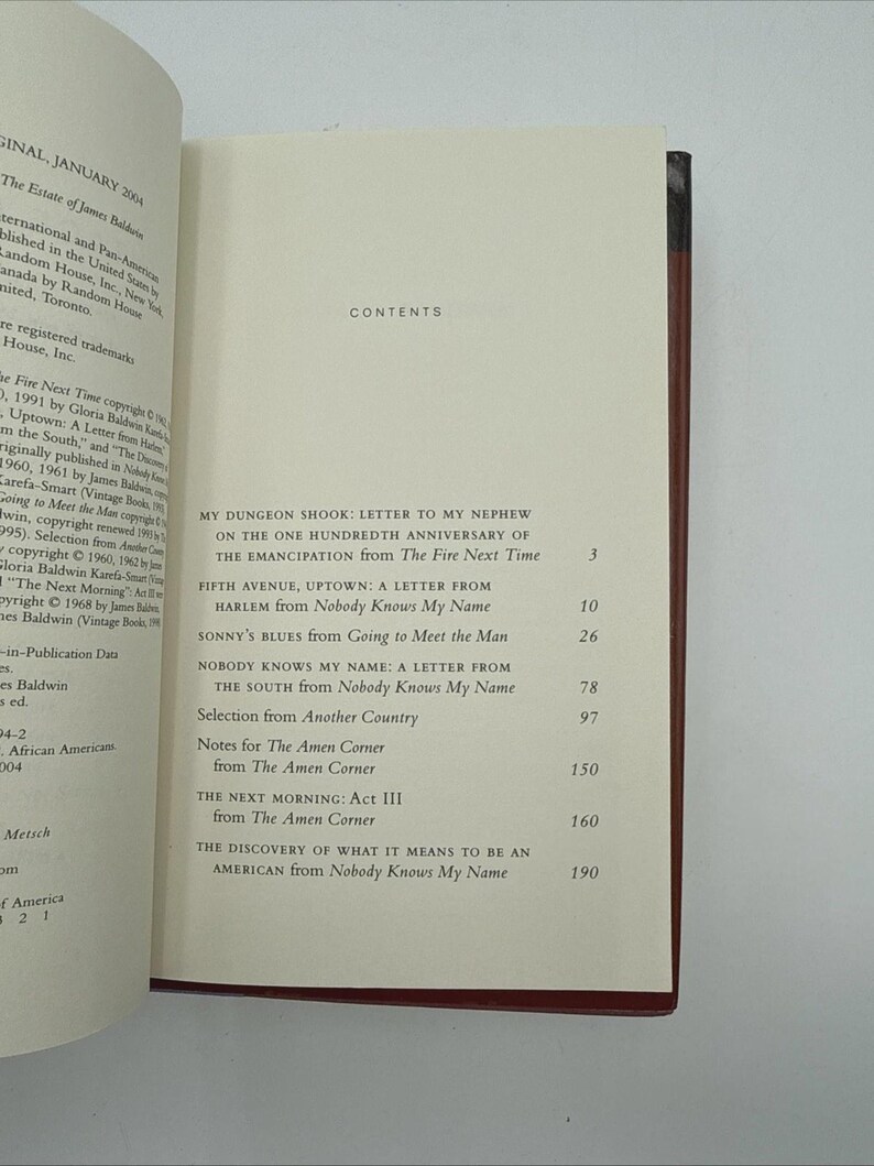 May include: Open book with a red spine, displaying the contents page of a book. The title "Nobody Knows My Name" is visible, along with other chapter titles. The book is open to a page with text.