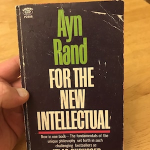 Pode incluir: Um livro de bolso preto e verde com o título "Ayn Rand Para O Novo Intelectual". A capa do livro apresenta o texto "Agora em um único livro - Os fundamentos da filosofia única expostos em best-sellers tão desafiadores como ATLAS SHRUGGED e A FONTE."
