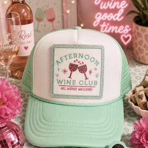 May include: A mint green and white trucker hat featuring a square patch with a silver glitter border. The patch displays the embroidered text "AFTERNOON WINE CLUB" and "ALL WINES WELCOME" with an illustration of two clinking wine glasses.