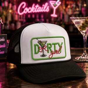 May include: A white and black trucker hat featuring a rectangular patch with a green border. The patch displays an embroidered martini glass with olives and the green and red text "DIRTY girl" centered on the front panel of the cap.