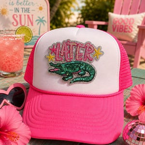 May include: A bright pink and white trucker hat featuring a sequined patch on the front panel. The patch displays a green alligator and the word LATER in pink letters, accented by two yellow stars and a silver glitter border.