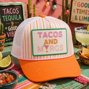 May include: A trucker hat featuring a white and orange striped front panel with a white mesh back. A rectangular patch on the front displays the embroidered text TACOS AND M RGS, with a small cocktail glass icon replacing the letter A.
