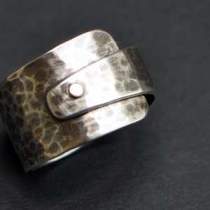 K&ouml;nnte beinhalten: Ein silberner Ring mit geh&auml;mmerter Textur und einem kleinen, runden Akzent aus Ros&eacute;gold. Der Ring ist so konzipiert, dass er am Finger getragen wird, wobei die geh&auml;mmerte Seite nach au&szlig;en zeigt.