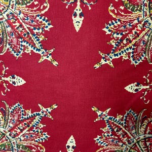 May include: Red fabric with a paisley pattern. The fabric has a textured surface and is decorated with a repeating pattern of intricate floral designs in shades of blue, green, and white.