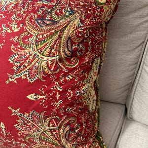 May include: Red paisley patterned throw pillow with a cream and green floral design. The pillow is shown on a cream coloured sofa.