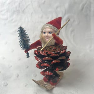 May include: A vintage Christmas ornament featuring a gnome figure atop a pinecone. The gnome wears a red velvet hat and jacket, holding a small tree and a gold-colored staff. The pinecone is painted with red accents, and the gnome is on skis.