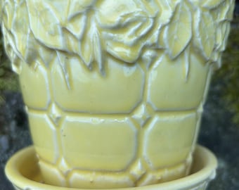 May include: A pale yellow ceramic plant pot with a matching saucer. The pot has a textured design of leaves and flowers around the rim and a geometric pattern on the body. The saucer is round and has a similar geometric pattern.