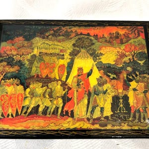 May include: A rectangular black lacquer box featuring a detailed hand-painted scene of a historical gathering with figures in robes and armor, a central yellow tent, and a distant city landscape, all framed by a thin gold decorative border.