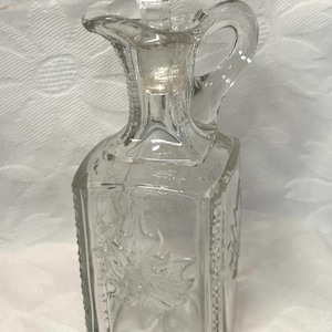 May include: Clear glass cruet with a square base, a handle, and a spout. The cruet features embossed floral designs on the front and back panels. It includes a glass stopper with a decorative top. The cruet is designed for serving oil or vinegar.