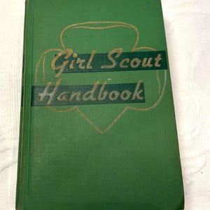 May include: A vintage green Girl Scout Handbook. The cover features the words "Girl Scout Handbook" in gold lettering and a gold outline of the Girl Scout emblem. The book is set against a white background.
