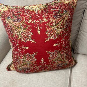 May include: Red cushion with a paisley pattern in shades of cream, green, and blue. The cushion is on a light grey sofa.