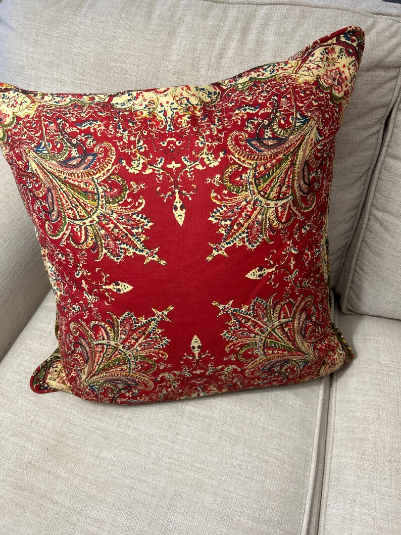 May include: Red cushion with a paisley pattern in shades of cream, green, and blue. The cushion is on a light grey sofa.