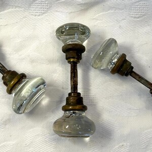 May include: Three antique glass doorknobs with brass hardware. The knobs are clear with a rounded shape, and the brass shows signs of age. The doorknobs are arranged on a white, textured surface.
