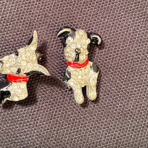 May include: A pair of black and white rhinestone dog pins with red collars. The dogs are facing different directions.