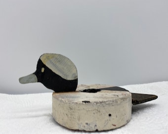 May include: A wooden duck decoy with a white, black, and gray painted design. The decoy is sitting on a white surface.