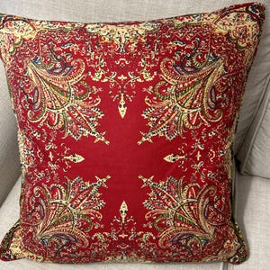 May include: Red cushion with a paisley pattern in shades of green, cream, and pink. The cushion is square shaped and has a soft, plush texture.
