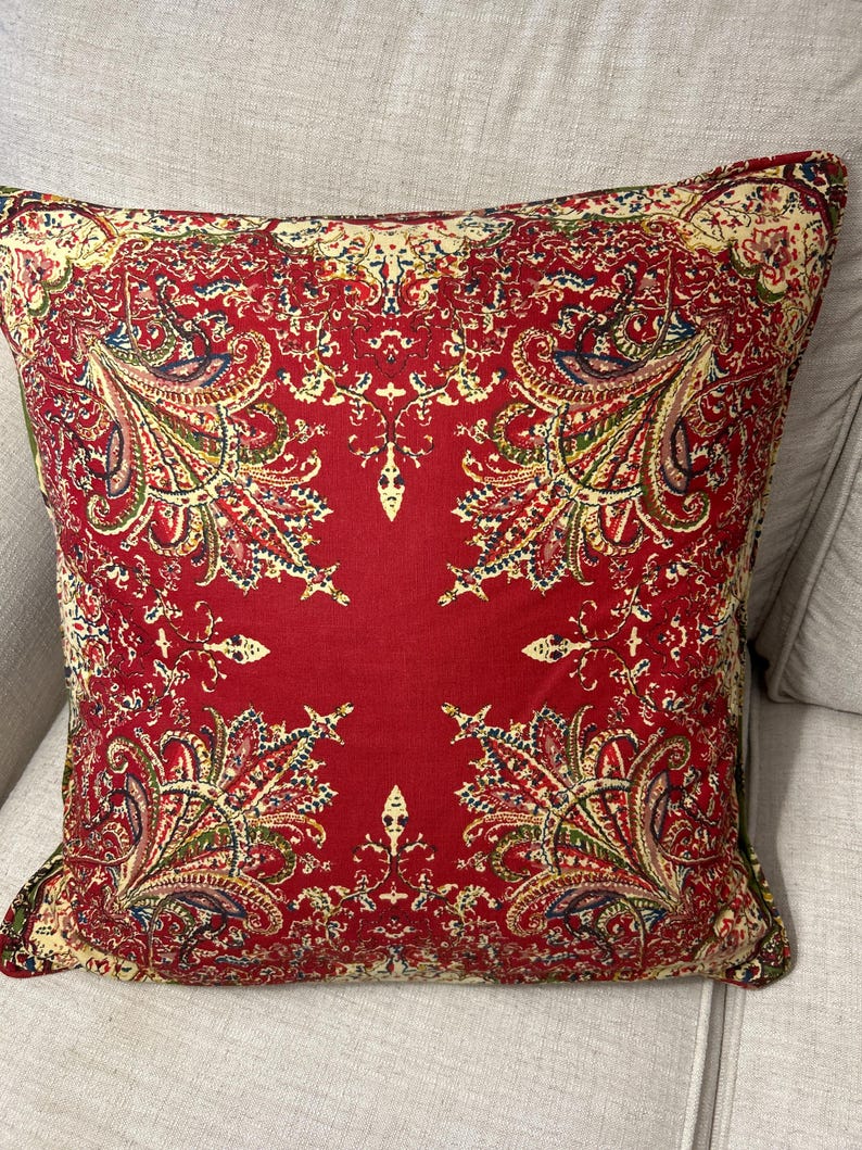 May include: Red cushion with a paisley pattern in shades of green, cream, and pink. The cushion is square shaped and has a soft, plush texture.