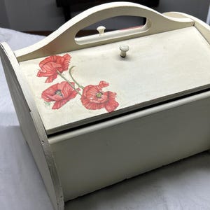 May include: A white wooden bread box with a handle and a floral design featuring red poppies. The box has a hinged lid and a white knob.