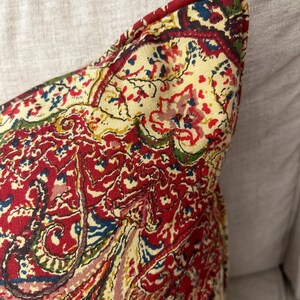 May include: A red, yellow, blue and green paisley patterned throw pillow with a cream background.