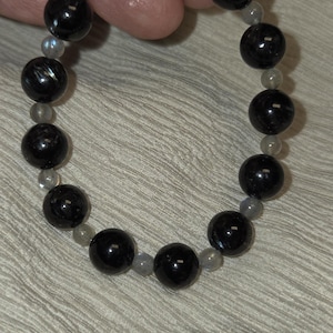 Peut inclure: Bracelet de perles composé de grosses perles noires brillantes alternant avec de plus petites perles grises. Les perles, d'environ 1 cm de diamètre, sont enfilées sur un cordon souple.