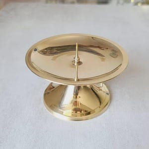 Pu&ograve; includere: Un portacandele color oro con una parte superiore rotonda e piatta e una base corta a piedistallo. Il portacandele ha una punta centrale per fissare una candela. La superficie &egrave; riflettente e il design &egrave; semplice ed elegante.