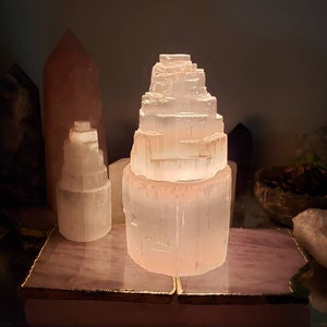 Op de afbeelding: Een witte selenietkristal lamp op een roze en gouden basis. De lamp is piramidevormig en wordt van binnenuit verlicht, waardoor een warme gloed ontstaat.