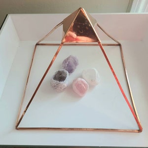 May include: A copper-toned metal pyramid with four sides and a square base. The pyramid is sitting on a white surface and contains three crystals: a purple amethyst, a pink rose quartz, and a clear quartz.