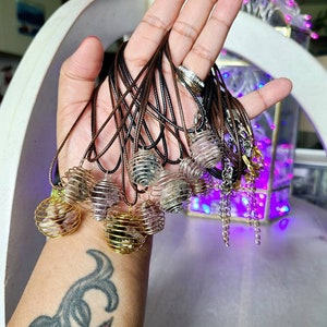 May include: A collection of crystal cage necklaces with various colored crystals. The necklaces are made with brown cord and gold metal cages. The cages are filled with crystals in shades of pink, purple, and clear.