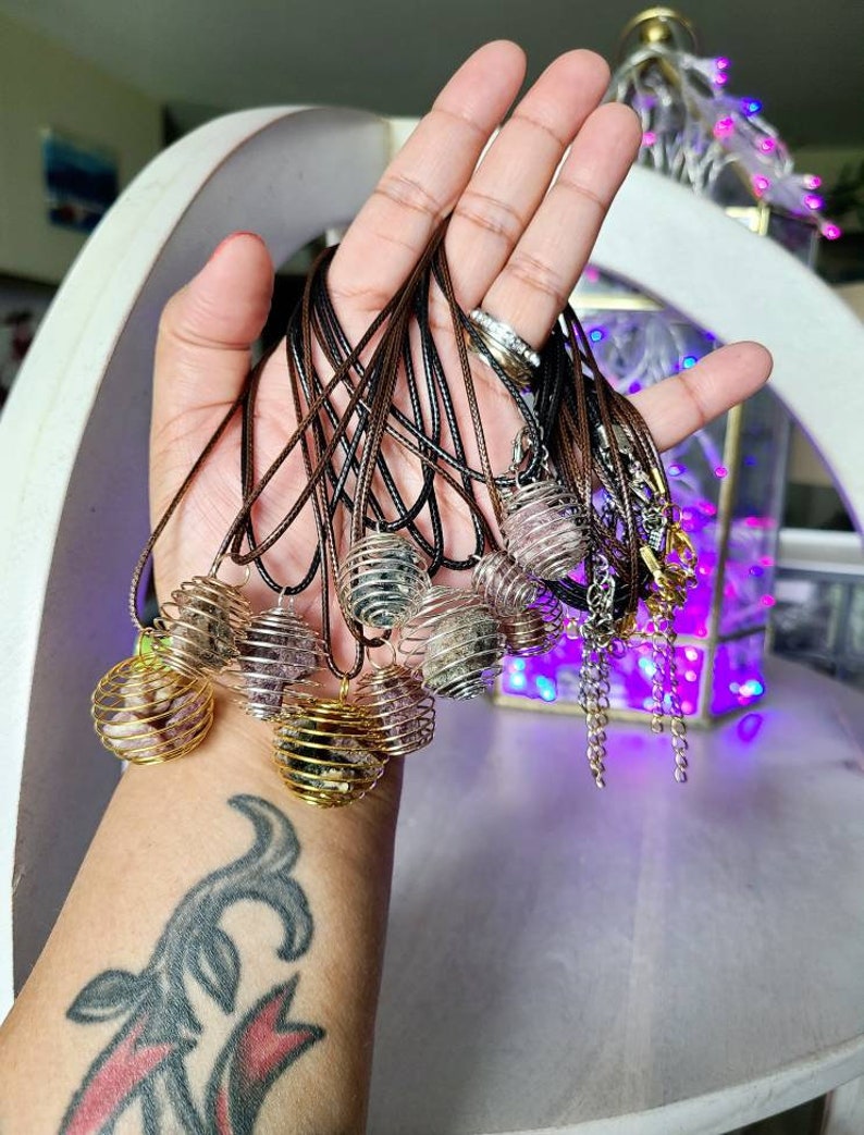 May include: A collection of crystal cage necklaces with various colored crystals. The necklaces are made with brown cord and gold metal cages. The cages are filled with crystals in shades of pink, purple, and clear.