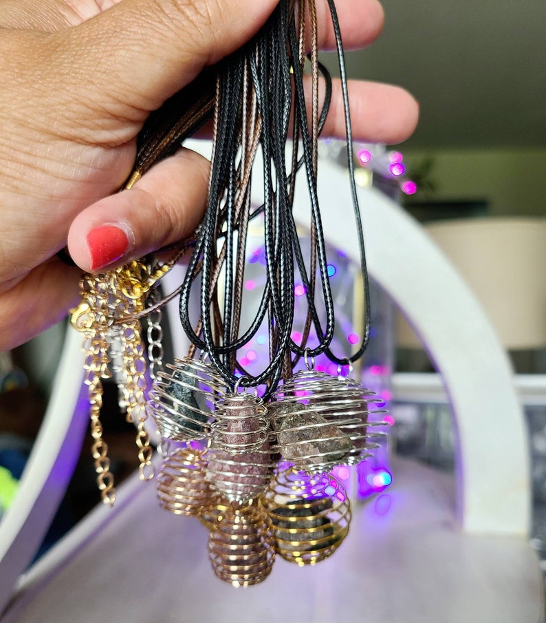 May include: A collection of necklaces with caged crystal pendants. The pendants are made of gold wire and contain various types of crystals, including amethyst, rose quartz, and black obsidian. The necklaces are strung on black and brown cords.