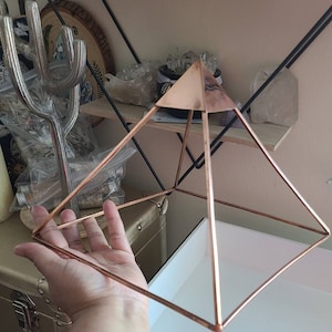 May include: A rose gold metal pyramid with a square base. The pyramid is made of thin wire and has a geometric design.