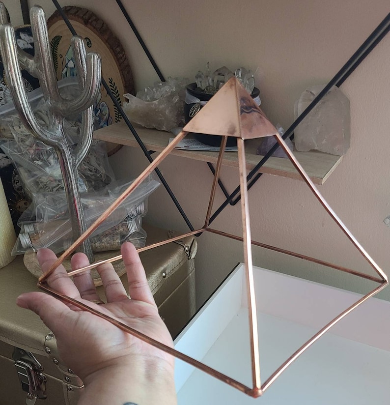 May include: A rose gold metal pyramid with a square base. The pyramid is made of thin wire and has a geometric design.