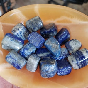 May include: A collection of tumbled lapis lazuli stones in a variety of shades of blue with white veining. The stones are arranged in a shallow orange bowl.