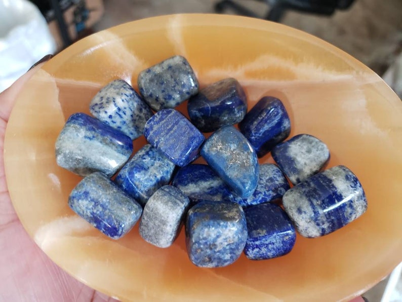 May include: A collection of tumbled lapis lazuli stones in a variety of shades of blue with white veining. The stones are arranged in a shallow orange bowl.