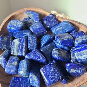 May include: A collection of tumbled lapis lazuli stones in a wooden bowl. The stones are a deep blue color with white veins running through them.