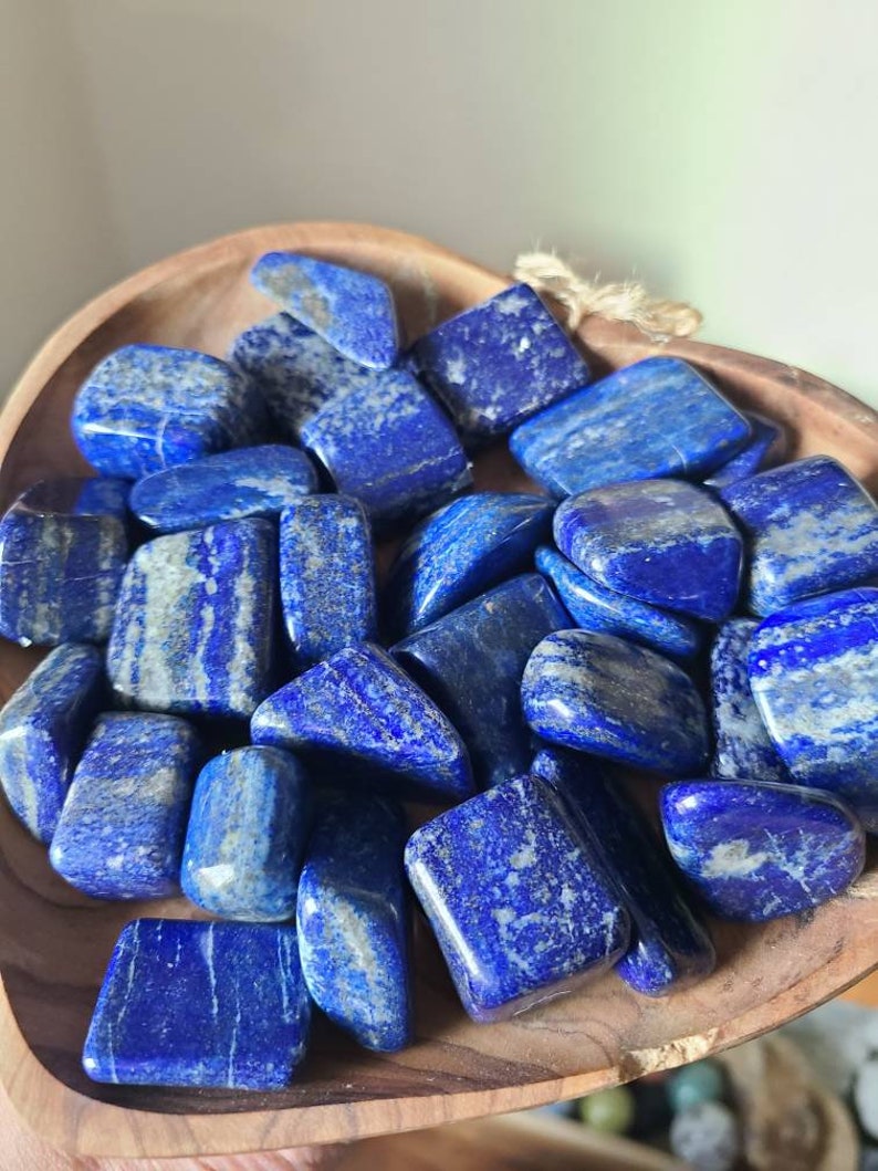 May include: A collection of tumbled lapis lazuli stones in a wooden bowl. The stones are a deep blue color with white veins running through them.